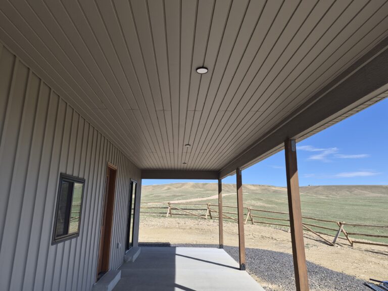 Covered porch with textured taupe metal soffit, wood posts, and open Wyoming hillside view beyond the home.