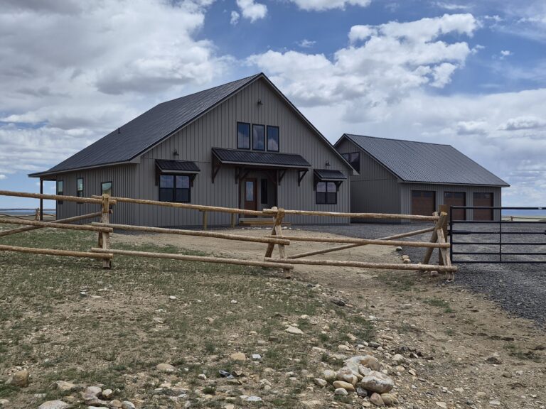 Custom home in Wyoming with textured taupe vertical metal siding, black roof, attached garage, and rustic fence in front.